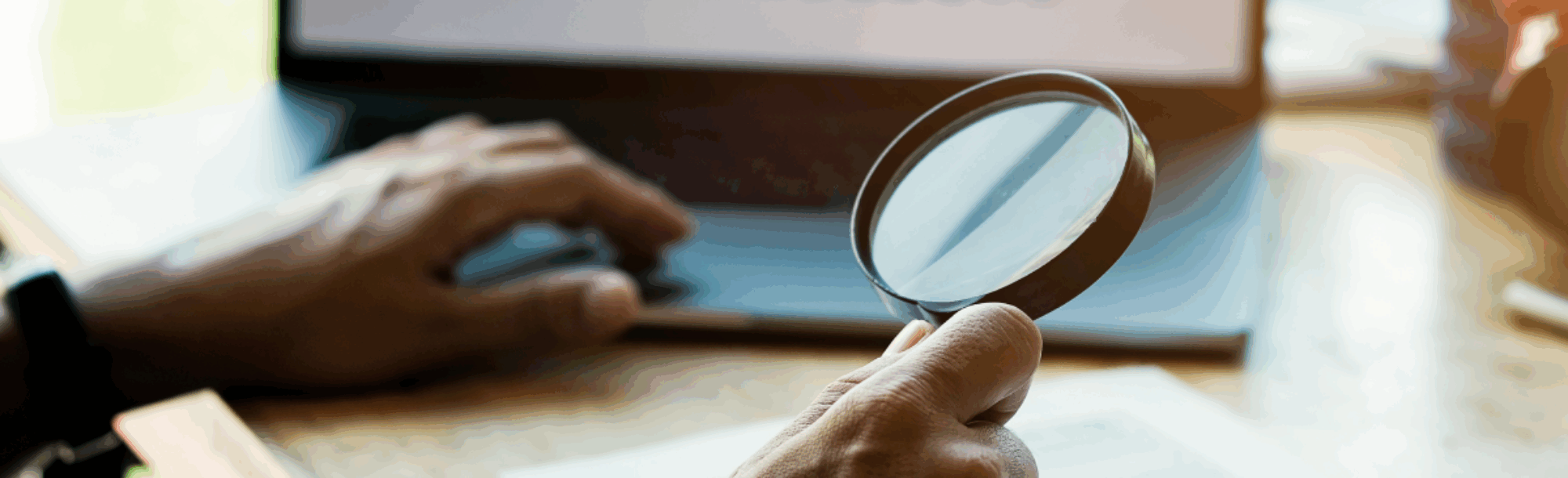 Set of hands--one hand holding a magnifying glass over a document, the other on a laptop keyboard. The laptop screen is displaying graphs.